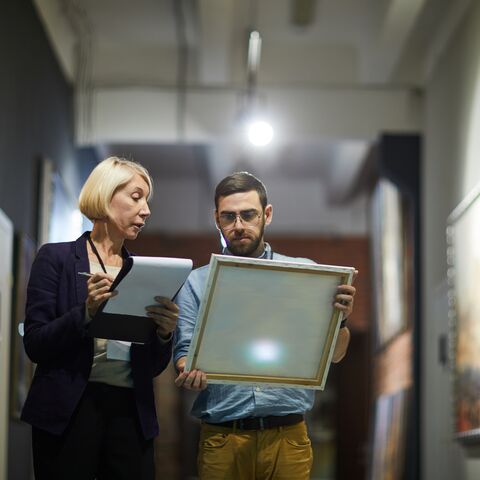 Eine Frau und ein Mann stehen in einer Gallerie mit alten Meistern an den Wänden. Er zeigt ihr ein Bild und sie macht sich Notizen.