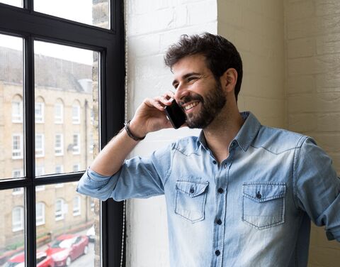 A smiling man in a light blue denim shirt talks on the phone next to a large window through which a brick façade and parked cars are visible.