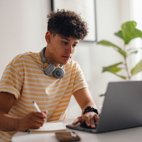 Un ragazzo con le cuffie al collo guarda il suo laptop e prende appunti.