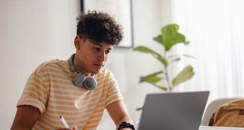 A teenager with headphones around his neck looks at his laptop and takes notes.