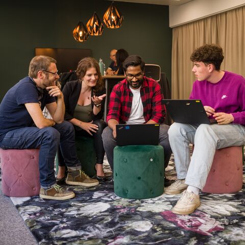 Four people sit on stools around a laptop and discuss in a cozy room with a dark wall and modern lighting.