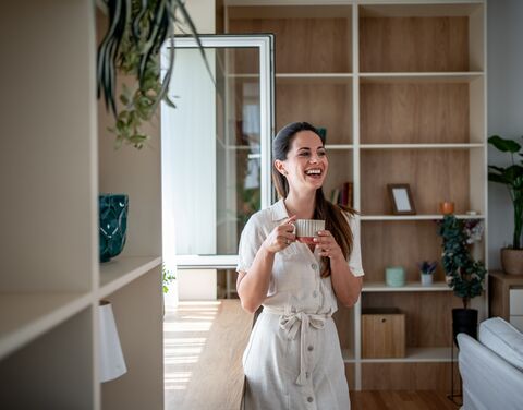 Junge Frau steht glücklich in ihrer modern eingerichteten Wohnung mit einer tasse in der Hand