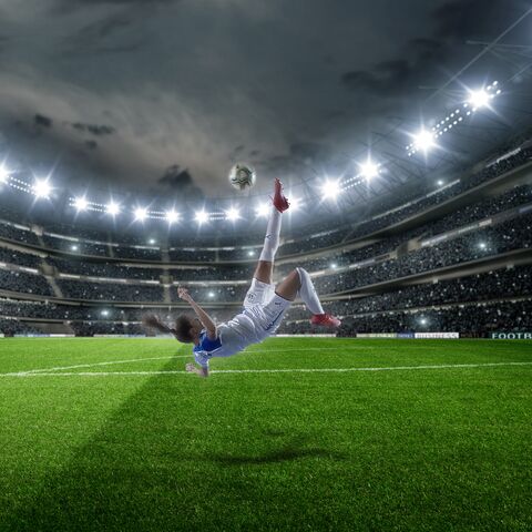 Eine Fussballspielerin macht einen spektakulären Fallrückzieher unter Flutlicht in einem vollbesetzten Stadion bei bewölktem Himmel.