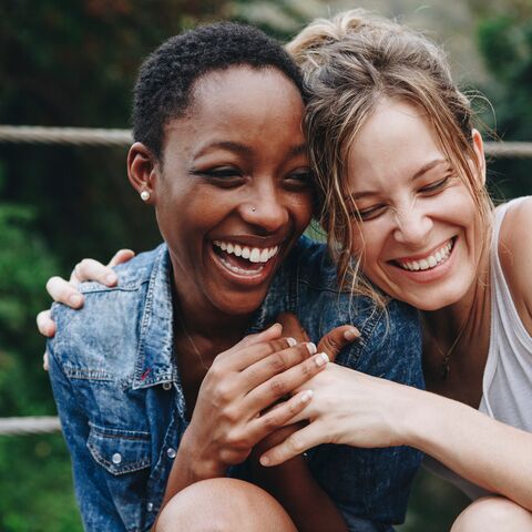 Deux jeunes femmes qui s'enlacent en riant ; l'une porte une veste en jean, l'autre un haut blanc. En arrière-plan, on aperçoit une nature verdoyante.