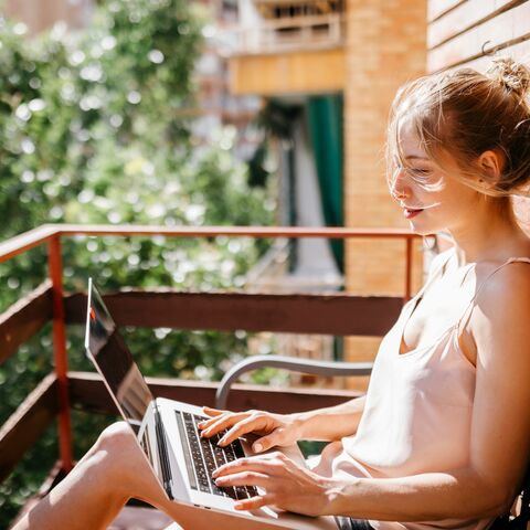 A young woman sitting on the balcony and looking relaxed at her laptop.