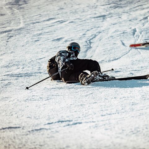 Un jeune homme en combinaison de ski est allongé dans la neige. Un ski est posé au-dessus de lui. Il tient un bâton de ski dans la main.