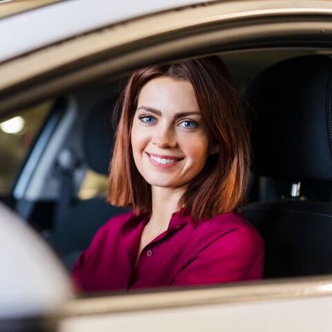 Eine Frau mit blauen Augen und roten Haaren lächelt aus dem Seitenfenster ihres Autos.