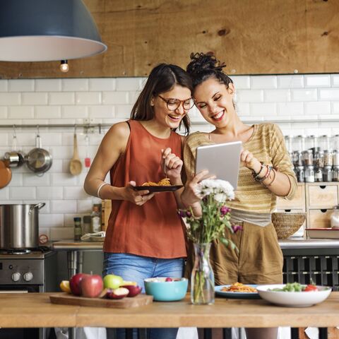Due donne che ridono sono in una cucina moderna, mangiano pasta e guardano insieme un tablet. Sul tavolo ci sono frutta e fiori.