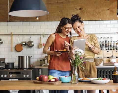 Zwei lachende Frauen stehen in einer modernen Küche, essen Pasta und schauen gemeinsam auf ein Tablet. 