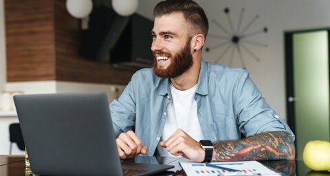 A man is sitting at a table smiling and working on his laptop.