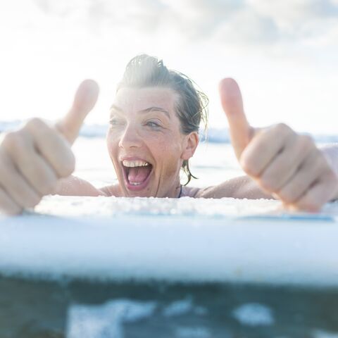 Una donna si aggrappa alla tavola da surf. Ride e tiene entrambi i pollici alzati.