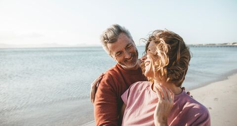 Ein älteres Paar umarmt sich lachend am Strand, im Hintergrund sieht man das Meer und einen klaren Himmel.