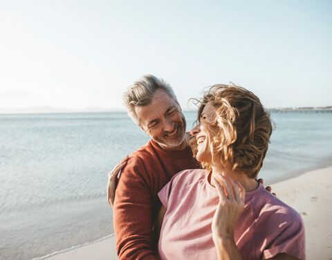 Ein älteres Paar umarmt sich lachend am Strand, im Hintergrund sieht man das Meer und einen klaren Himmel.