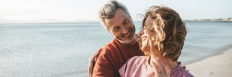 An older couple laughingly hugging on the beach, in the background you can see the sea and a clear sky.
