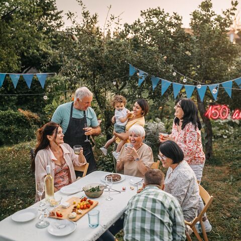 Des membres d’une famille sont assis ou debout autour d’une table couverte d’une nappe blanche dans un jardin. Ils rient et font la fête. Une guirlande lumineuse et des fanions sont suspendus au-dessus d’eux.