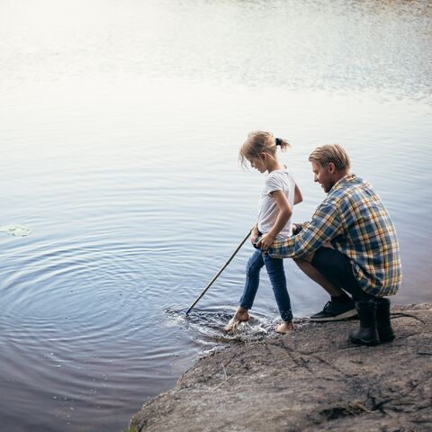 Un homme et un enfant se tiennent au bord d'un lac, l'enfant tient un bâton dans l'eau et l'homme s'agenouille à côté de lui et le soutient.