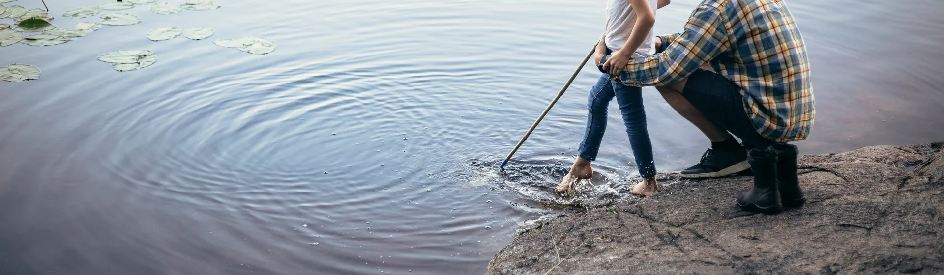 Ein Mann und ein Kind stehen am Ufer eines Sees, das Kind hält einen Stock ins Wasser und der Mann kniet neben ihm und unterstützt es.