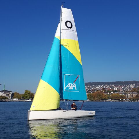 A sailboat with a yellow, blue, white sail is cruising in front of a city.