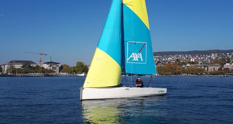 A sailboat with a yellow, blue, white sail is cruising in front of a city.