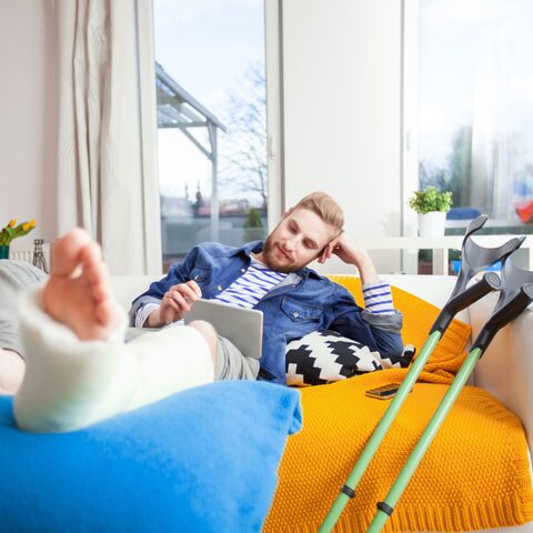 Un homme avec une jambe dans le plâtre est allongé sur un canapé, entouré de béquilles et de coussins, et utilise une tablette dans un salon lumineux et confortable.