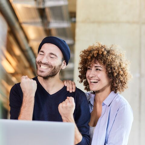 Ein Mann und eine Frau lächeln und jubeln gemeinsam vor einem Laptop in einem modernen, gut beleuchteten Büro.