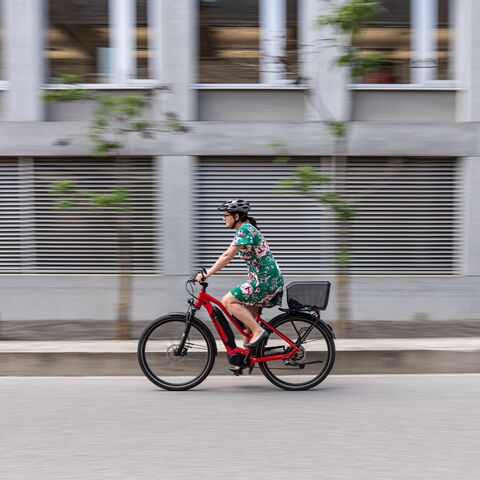 Donna in sella a una e-bike rossa e abito a fiori verdi passa accanto a un edificio grigio.