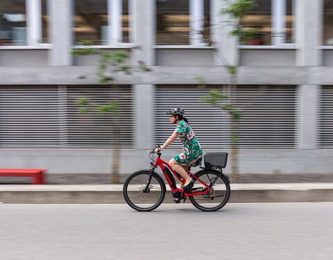 Une femme portant un casque de vélo et une robe à fleurs vertes passe devant un bâtiment gris sur un vélo électrique rouge.