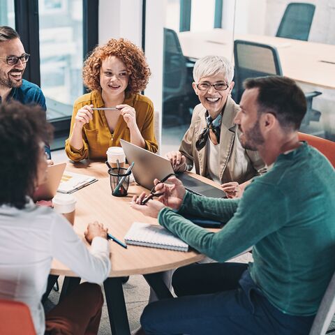 Un gruppo di cinque persone siede intorno a un tavolo rotondo in un ufficio moderno, discutendo allegramente mentre prende appunti e usa il computer portatile.