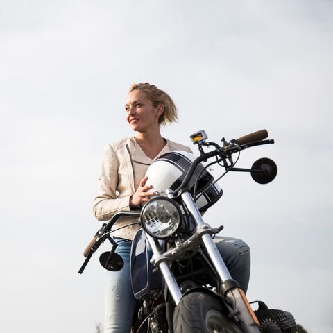Une jeune femme aux cheveux blonds, souriante, est assise sur une moto en plein air, sous un ciel nuageux, et tient son casque à la main.