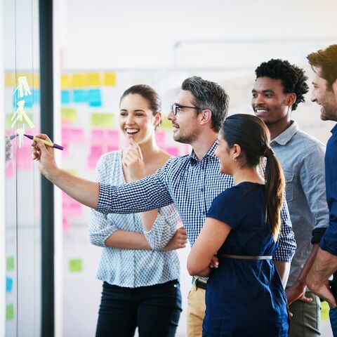 Eine Gruppe von fünf Personen steht vor einem Whiteboard, lächelt und diskutiert, während eine Person darauf zeichnet.