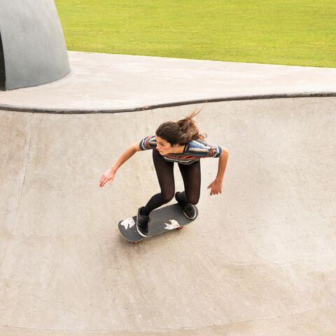 A young woman in casual clothes rides a skateboard in a skate park while taking a turn in a half-pipe.