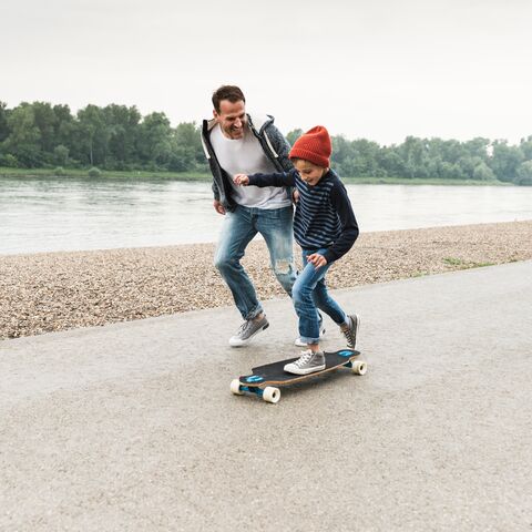 Un garçon fait du skateboard et son père lui tient la main. À l’arrière-plan, on voit une rivière.