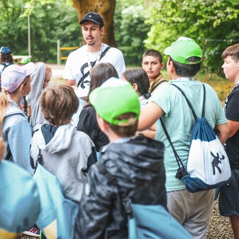 A man explains something to a group of children outdoors. The children are wearing backpacks and caps.