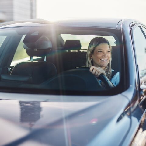 A smiling woman with blonde hair drives a car and wears a yellow sweater while looking through the open window.