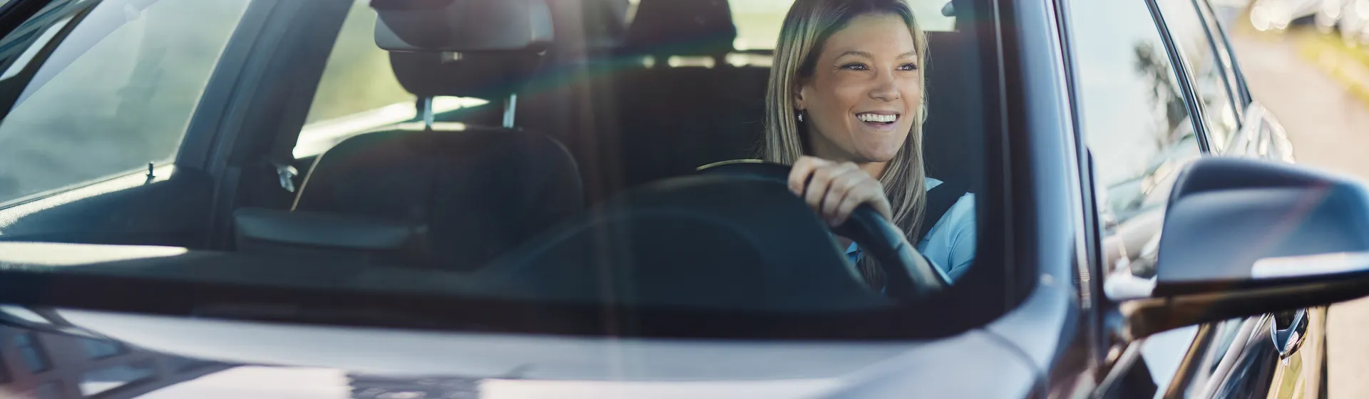 A smiling woman with blonde hair drives a car and wears a yellow sweater while looking through the open window.
