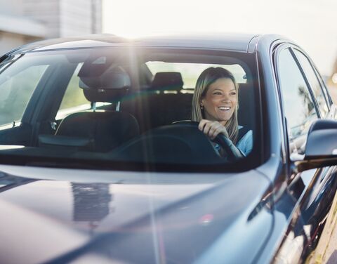 A smiling woman with blonde hair drives a car and wears a yellow sweater while looking through the open window.