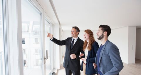 A young couple is standing in an empty apartment together with a real estate agent. All three are smiling and looking out a window wall.