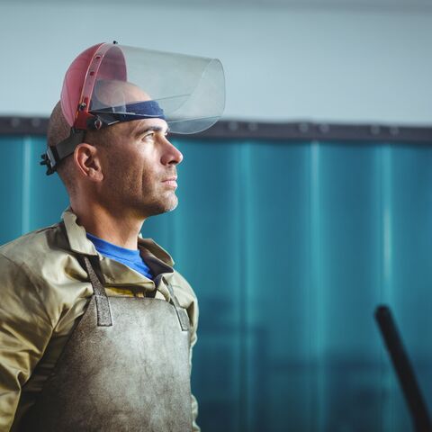 Un homme vêtu de vêtements de protection et d'un écran facial se tient dans un atelier, le regard concentré vers l'avant, devant un rideau bleu.
