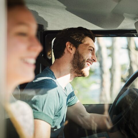 Un homme souriant conduit une voiture, tandis qu'une femme souriante est assise sur le siège du passager. Le soleil brille à travers la fenêtre.