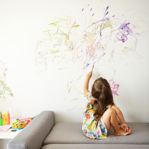 Ein kleines Kind sitzt auf einem grauen Sofa und malt mit bunten Farben auf eine weisse Wand, neben ihm liegt Malzubehör auf einem Tisch.