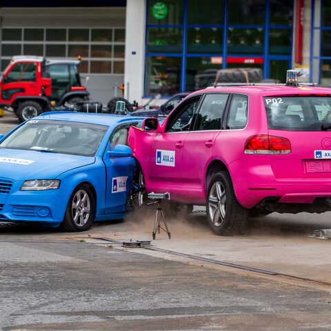 Un SUV rosa urta lateralmente un’auto blu, entrambi i veicoli recano il logo AXA e sono parte di un crash test effettuato presso un circuito di test.