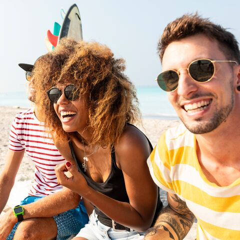 Una giovane donna e due giovani uomini seduti sulla spiaggia ridendo. Sullo sfondo ci sono tavole da surf e si vede l’oceano.