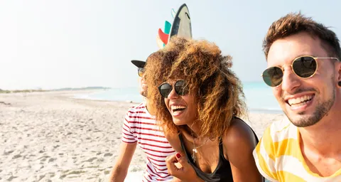 Une jeune femme et deux jeunes hommes sont assis sur la plage en riant. À l’arrière-plan, il y a des planches de surf et on voit la mer.