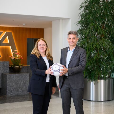 Two people in business clothes are holding a soccer ball together in a modern office with the AXA logo in the background and a large plant.