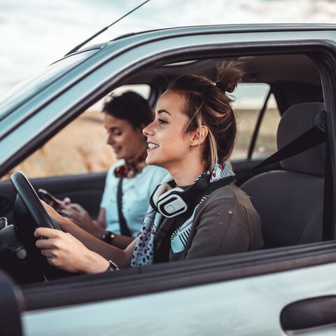 Zwei Freundinnen fahren fröhlich in die Ferien. Die eine schaut lächelnd, aber konzentriert voraus, während ihre Freundin auf dem Handy die nächste Destination eingibt.