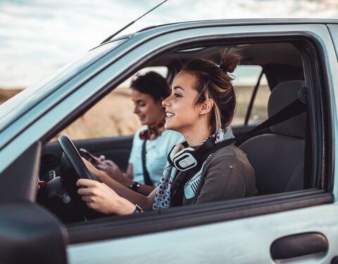 Zwei Freundinnen fahren fröhlich in die Ferien. Die eine schaut lächelnd, aber konzentriert voraus, während ihre Freundin auf dem Handy die nächste Destination eingibt.
