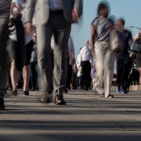 Une foule en tenue de travail marche en hâte sur un trottoir dans une ville, les jambes et les pieds des passants bougent vaguement.