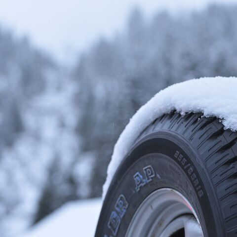 A snow-covered car tire in the foreground, a wintery, snow-covered forest landscape in the background.