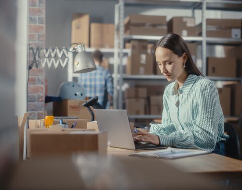 A woman is working intently on her laptop, with a warehouse visible in the background.