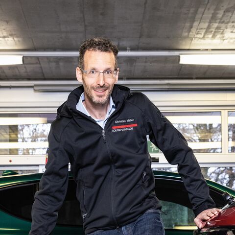 Ein lächelnder Mann in einer schwarzen Jacke steht in einer Garage zwischen zwei Autos, einem grünen und einem roten, vor einer Fensterfront.
